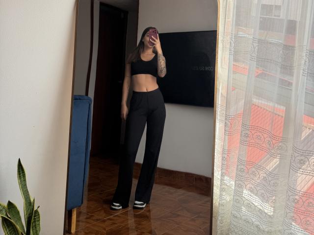 ThaliaAres with tattoos, black crop top, black pants, and white shoes, standing in front of mirror, holding phone, showing slim, toned midriff, and small ass.