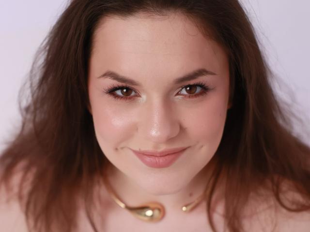 Dreia has brown eyes, long brown hair, a slight smile, and wears a gold necklace. Her skin is fair with a subtle pink blush. She looks directly at the camera.