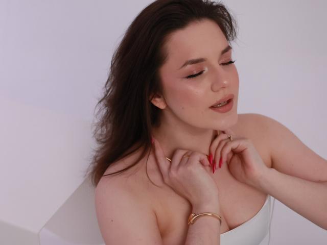 Dreia with closed eyes, dark brown hair, and pink lipstick, touching her neck with red-manicured fingers, wearing a white strapless top, and gold bracelets.