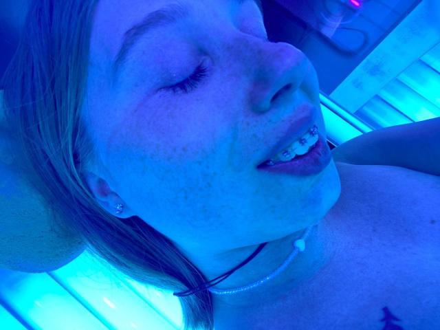 OuidaMcgrane's face in blue light, freckles, tongue out with a tooth piercing, short blonde hair, earrings, and necklace, lying down, showing her bare shoulders and a bit of her chest.