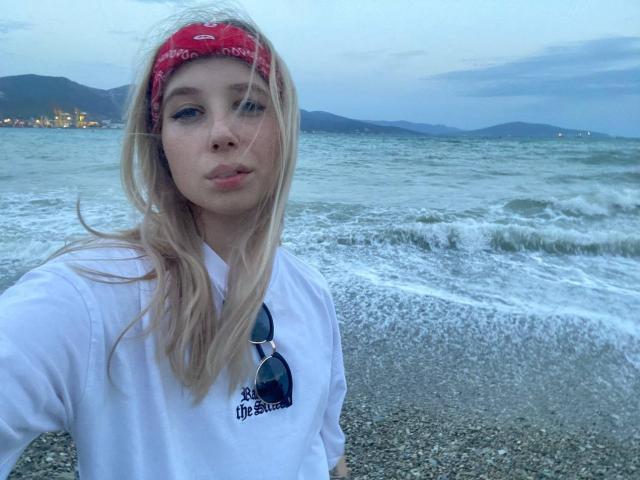 Blonde OuidaMcgrane with red bandana, white shirt, and sunglasses on chest, pouting at beach, water in background, mountains and city lights visible. Her tits are small.