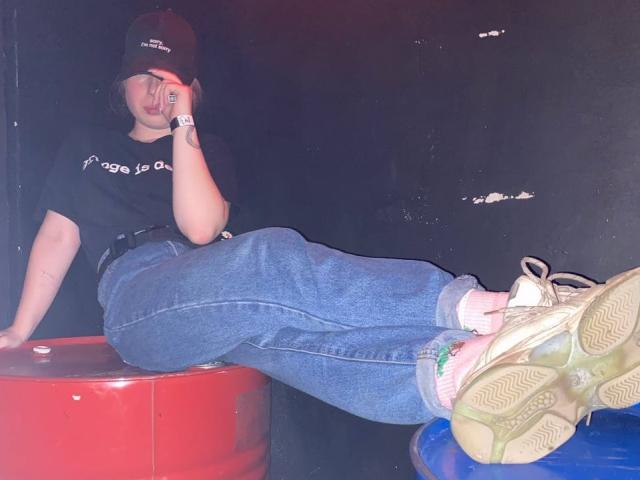 OuidaMcgrane sitting on a red and blue cylinder, wearing a black cap, black t-shirt, blue jeans, white sneakers, and a wristwatch, with her hand on her face.