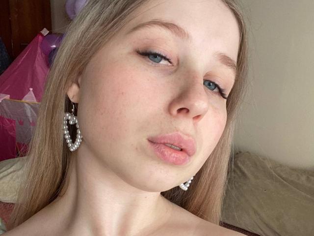 OuidaMcgrane has pale skin, blue eyes, and pink pouty lips. She's wearing pearl earrings and has blonde hair. Her eyelashes are long and she's giving a slight pout.