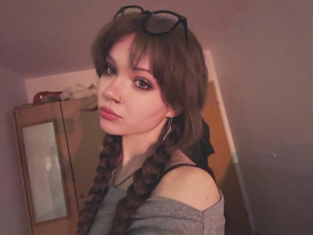 FannyEnsell has brown hair in braids, wearing a gray off-shoulder top, glasses on her head, and hoop earrings, with a nose ring, in a dimly lit room.