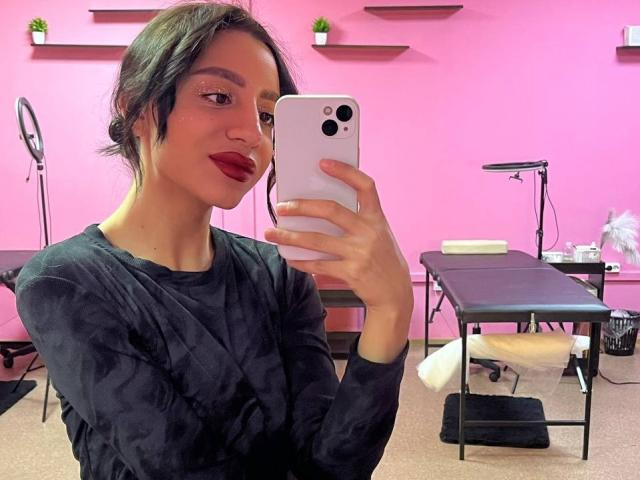 CharleenOachs with short black hair, wearing a black long-sleeve shirt, taking a selfie in a pink room, holding a white phone, with red lipstick and a slight smile.