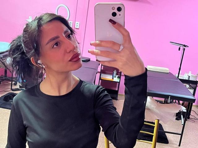 CharleenOachs with pouty lips, wearing a black long-sleeve top, holding a white iPhone, taking a mirror selfie, pink room, ring on her finger, and dark red nails.