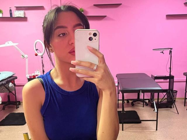 CharleenOachs with dark hair, wearing a blue sleeveless top, taking a mirror selfie in a pink room with medical equipment, holding a white iPhone, looking at the camera.