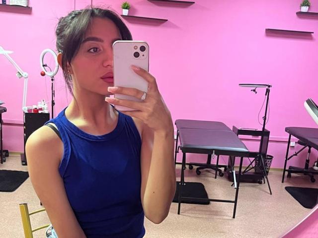 CharleenOachs with medium tits, wearing a blue tank top, taking a mirror selfie in a pink room with exercise equipment, holding a white iPhone, has dark hair and red lips.