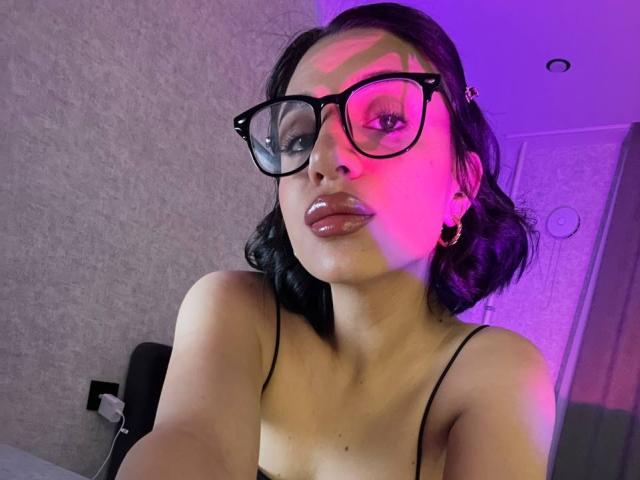 CharleenOachs with black glasses, pinkish-purple light on her face, medium tits, shiny pink lips, black hair, and gold hoop earrings, in a selfie.