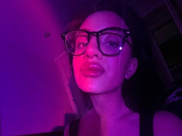 CharleenOachs with shiny pink lips, wearing black glasses, and dark hair, lit in purple and pink neon, showing a confident smile. Her tits are partially visible in the frame.