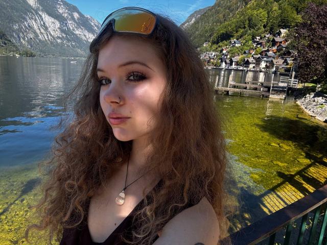 FannyEnsell with curly brown hair, wearing sunglasses on her head, black top, necklace with seashell, standing by a clear, green-tinted lake with wooden docks and mountains in the background.