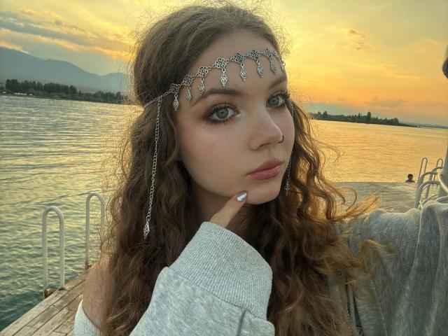 FannyEnsell with curly brown hair, wearing a silver head chain, gray sweatshirt, nose ring, and silver earrings, pointing at her chin, near a lake at sunset.
