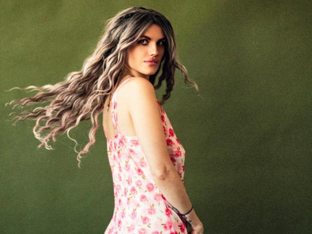 MilaLoona with long, wavy brown hair, wearing a pink floral dress, looking over her shoulder, green background, showing her ass and a black bracelet on her right wrist.