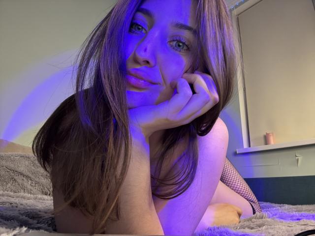 KitKristy with long brown hair, blue-tinted face, and fishnet dress, laying on bed, smiling, resting chin on hand, showing off her tits, with a relaxed, sexy expression.