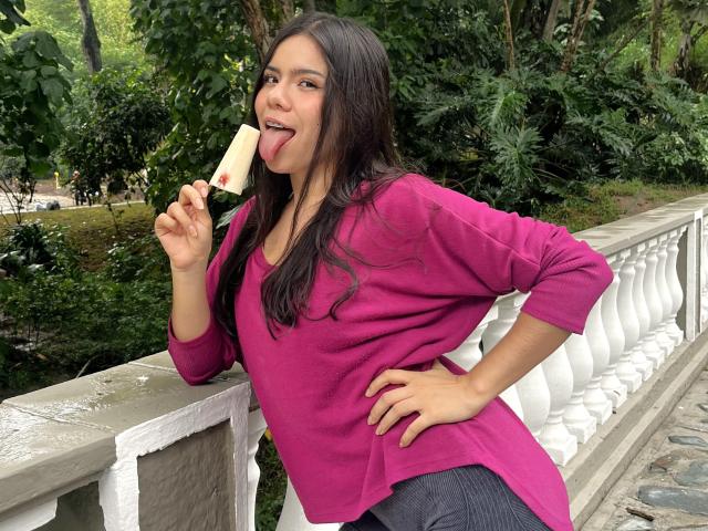 MaraCortes, with long brown hair, pink shirt, and black pants, leans on a white railing, tongue out, eating a white popsicle with red dot, looking playful.