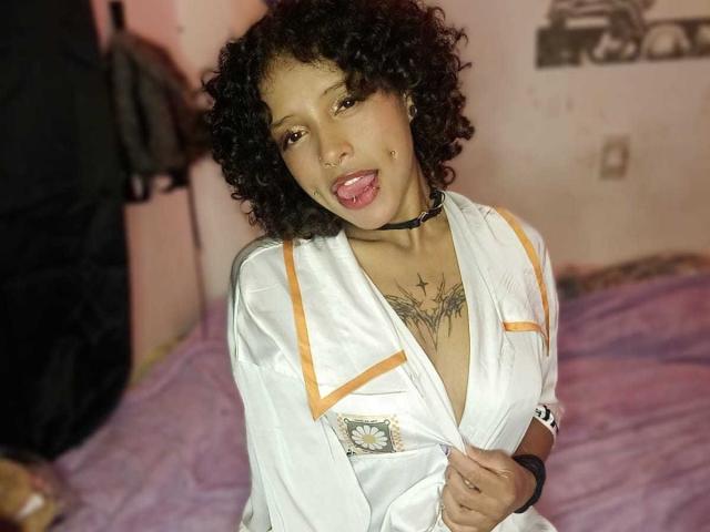 LunaKeller with curly black hair, tan skin, tongue out, wearing a white satin robe with orange trim, black choker, and a star tattoo on her chest. Boobs visible.