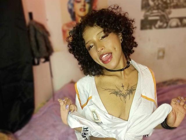 LunaKeller, com cabelo preto encaracolado, pele morena e seios pequenos, vestindo uma jaqueta branca, língua para fora, colar preto e tatuagem no peito, segurando a jaqueta aberta em um quarto..