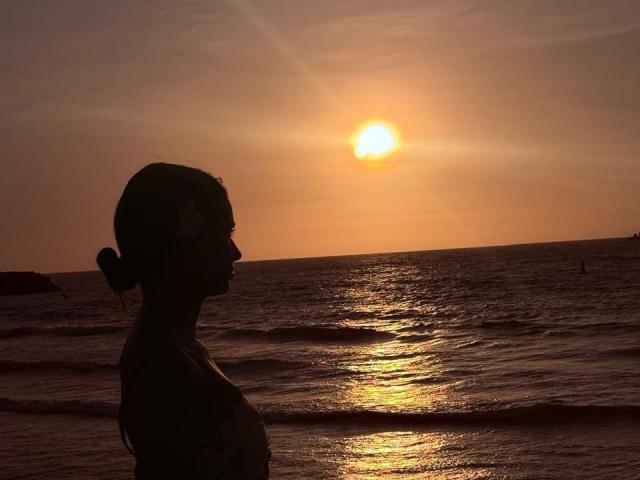 Silhouetted KimmyK with tits showing, hair in bun, against sunset over ocean, reflection on water, sun high, KimmyK's profile, water's edge, gentle waves, calm evening.
