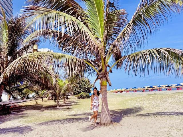 KimmyK stands by a tall palm tree with green and brown fronds, wearing a white floral dress, on a sunny beach with blue sky and colorful umbrellas in background.