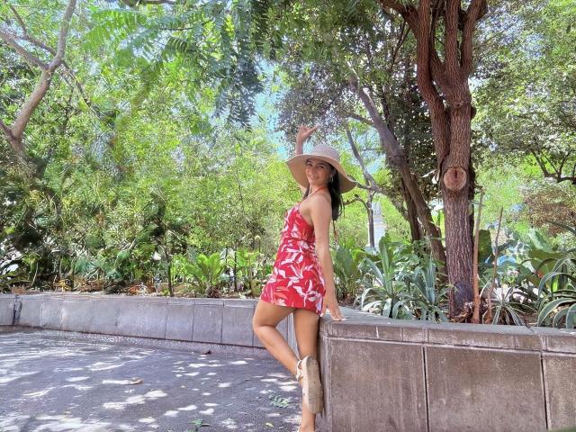 KimmyK with tan skin, long brown hair, red floral dress, straw hat, and tan boots, leaning on a concrete wall, trees and greenery in the background, sunny day.