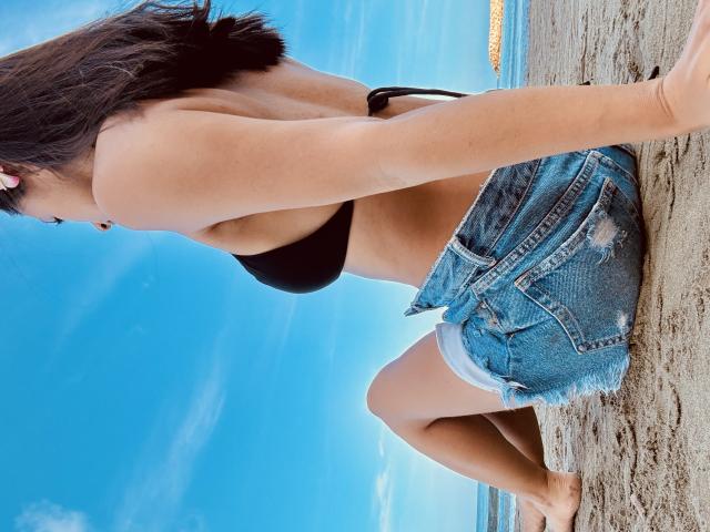 KimmyK with black bikini top, showing her boobs, and blue denim shorts, kneeling on sandy beach, barefoot, brown hair, pink earbuds, clear blue sky, and ocean in background.