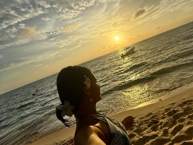 KimmyK with dark hair, white flower, wearing a green and white bikini, sits on sandy beach, back to camera, looking at sunset over ocean, sun reflecting on water.