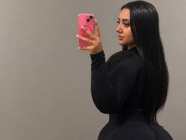 AmaliaDalton with long black hair, wearing a black long-sleeve top, holding a pink phone, taking a mirror selfie, showing her curvy butt and small tits.