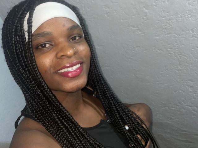 RenataWalker with dark skin, long black braids, white headband, black tank top, and pink glossy lips, smiling at the camera. Her tits are small, and her pussy is unseen.