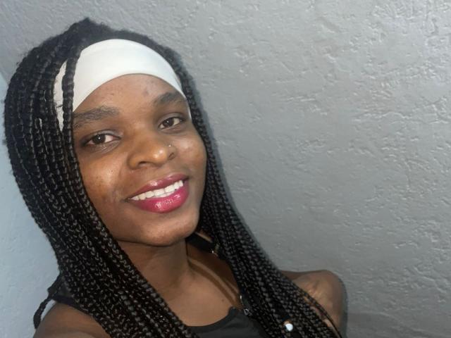 RenataWalker with dark skin, long black braids, white headband, black top, pink lipstick, smiling, small nose piercing, textured gray wall in background.