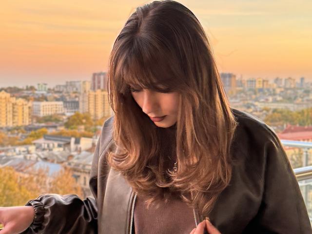 Adelisha with long brown hair, looking down, wearing a black leather jacket and brown top, standing on a balcony, sunset in the background, hands near her chest.