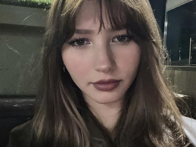 Adelisha has fair skin, brown eyes, and long brown hair with bangs. She's looking at the camera with a neutral expression, wearing dark lipstick. She's sitting in a dimly lit room.