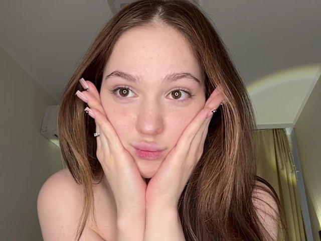 BaoRoebke has brown eyes, long brown hair, and pink lips. Her hands are touching her face with fingers spread. She's wearing silver earrings and has pink nails.