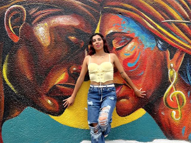 Carla Torres, con cabello castaño oscuro, una camiseta amarilla de encaje con corte y jeans azules rotos, apoyada contra una pared llena de grafitis coloridos que presenta una pintura facial grande y detallada en rojo y marrón..