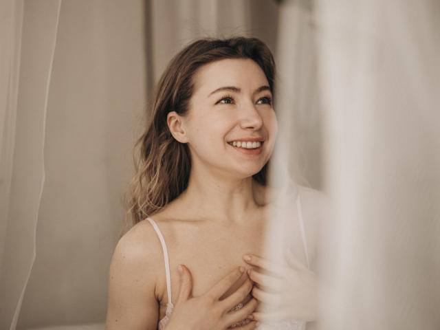 MiaFlirt with wavy brown hair, smiling, covering her small tits with her hands, wearing a white lace top, soft light, semi-transparent curtain in the foreground.