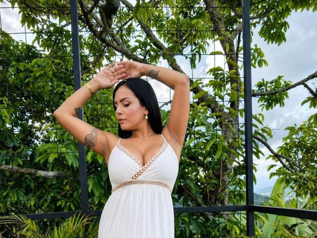 JessicaFiore with arms raised, wearing a white dress with lace trim, showing her tits and tattoos, black hair, standing in front of a metal fence with greenery.