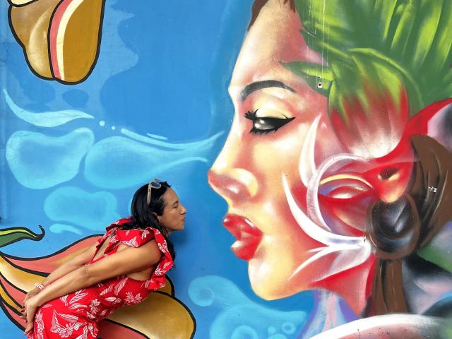 JadeFoster, with red floral dress and black hair, leans against giant, colorful, painted face with green hair and red lips, looking up. Boobs, ass, and face are detailed.