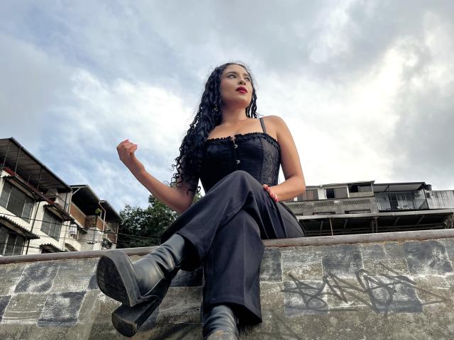 LilithStygian with curly black hair, red lipstick, black lace corset, black pants, and black boots, sitting on a stone ledge, legs crossed, looking up, cloudy sky background.