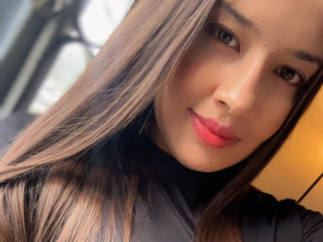 EmmaLombardi's face with long brown hair, brown eyes, and pink lipstick, wearing a black top, looking at the camera with a slight smile. Her tits are not visible.