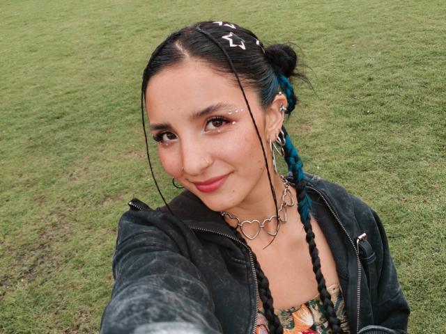 ArisSuarez with blue braids, heart choker, nose ring, and eyeliner, smiling in a selfie, wearing a black jacket, grassy background, showing her tits with a tattoo.