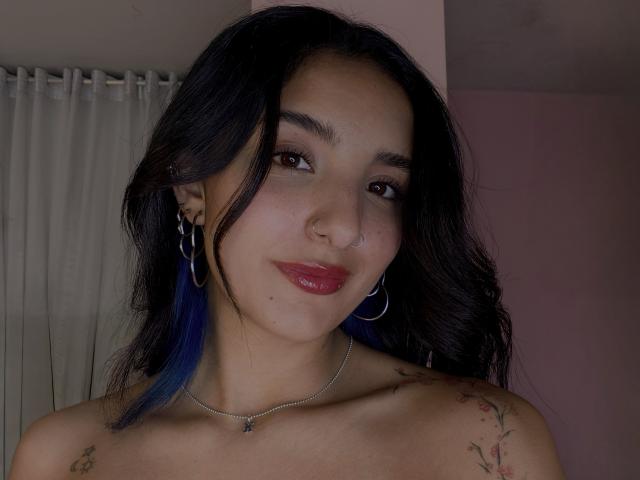 ArisSuarez with dark, wavy hair, blue tips, hoop earrings, nose ring, red lipstick, tattoos on shoulders, and a silver necklace, smiling softly.