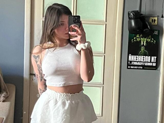 Mettis, avec de longs cheveux bruns, un crop top blanc et une jupe blanche, tenant un téléphone pour un selfie devant un miroir, montrant des tatouages sur le bras gauche, un élastique blanc au poignet droit, tenant un téléphone noir..