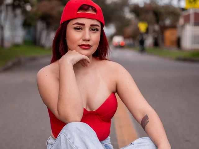 JulietaDupont with red hair, wearing a red cap, red strapless top, and blue jeans, sitting on a road, hand on chin, showing her tits and tattoo on left arm.
