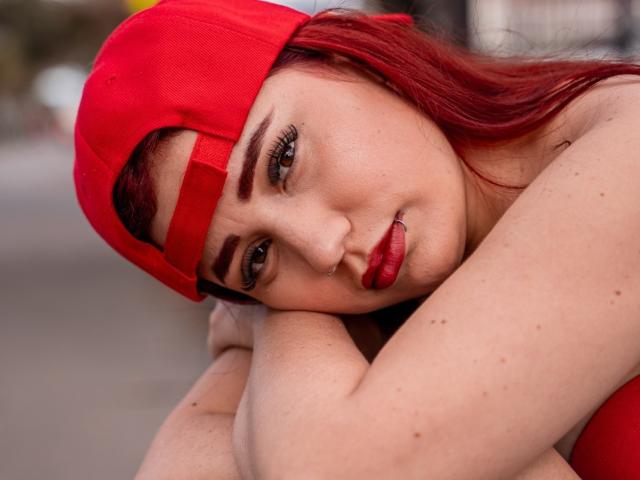 JulietaDupont with red hair, red cap, and red lipstick, resting her head on her arm, showing her tits, with a subtle nose ring and freckles, in a street shot.