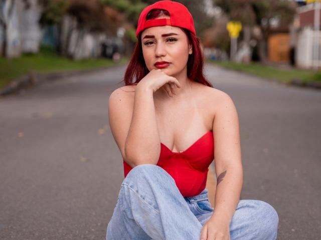 JulietaDupont with red hair, red tube top showing her tits, and blue jeans, sitting on a road, hand on chin, wearing a red cap, looking at the camera.