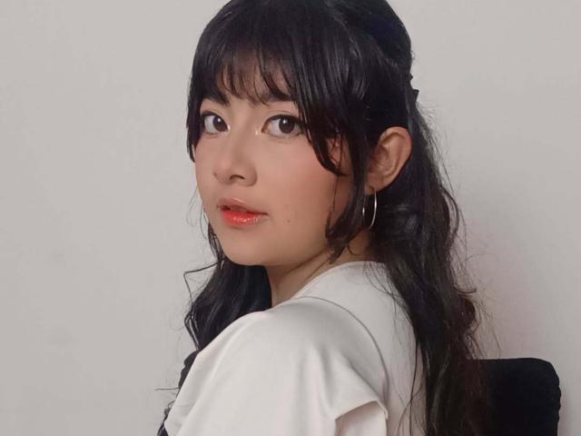 NekoPam with black wavy hair, bangs, and red lipstick, wearing a white shirt, looking over her shoulder, small hoop earrings, soft light on her face, slight smile.