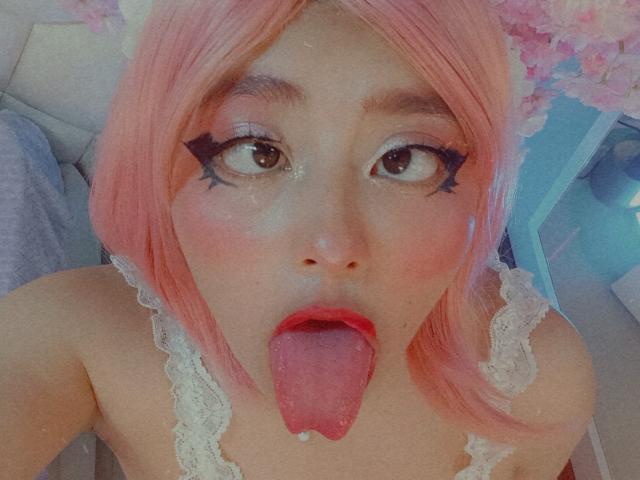 NekoPam with pink hair, big eyes, black star eyeliner, and tongue out. She's wearing a white lace top, and her tongue is slightly wet. NekoPam's face is centered.