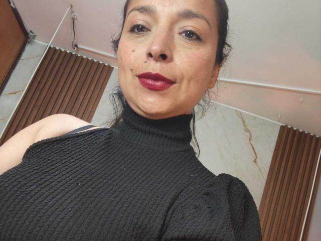 StangelarRivera with dark red lipstick, black textured turtleneck, small moles on face, dark hair in a bun, smiling, showing bare shoulders, standing in a simple room.