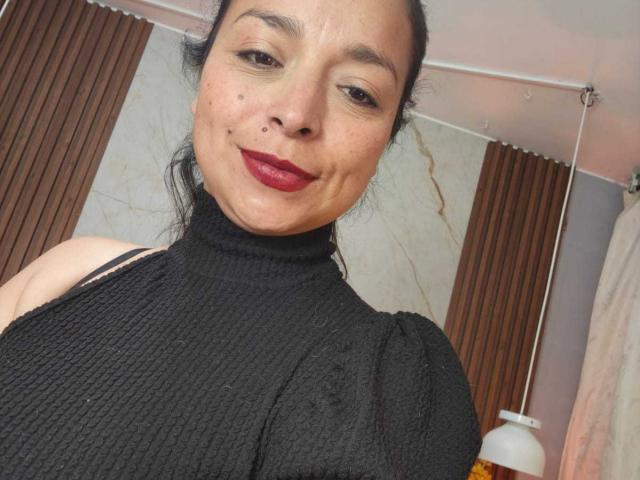 StangelarRivera with dark red lipstick, black textured turtleneck, and a small mole on her right cheek, smiling at the camera. Her black hair is tied back.