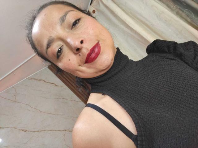 StangelarRivera has medium skin, dark hair in a bun, red lipstick, black textured top with strap showing, and a slight smirk. Her tits are small, visible through top.