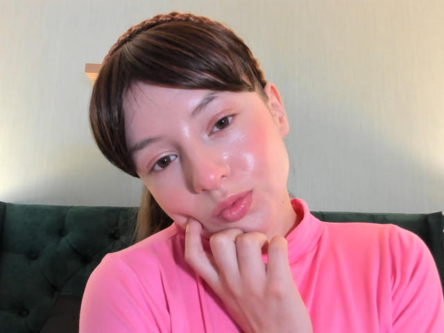 VioletaNova con piel brillante, labios rosados y gruesos, cabello castaño en una diadema, vistiendo una camisa rosa, con la mano debajo del mentón, mirando a la cámara, iluminación suave, fondo verde con textura..
